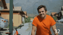a man in an orange shirt stands next to a red bicycle with the number 32 on the bottom right