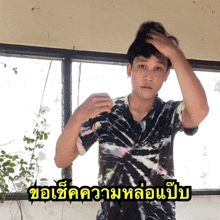 a man in a tie dye shirt holds his hand to his head in front of a window with a caption in thai