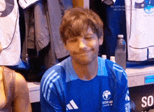 a man wearing a blue soccer aid shirt smiles in a locker room