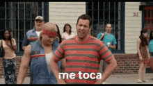 a man in a red and black striped shirt stands in front of a group of people with the words me toca on his chest