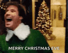 a man in a green sweater is laughing in front of a christmas tree and says merry christmas eve .