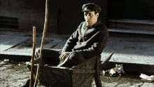 a man in a hat sits in a wheelbarrow on the side of the road
