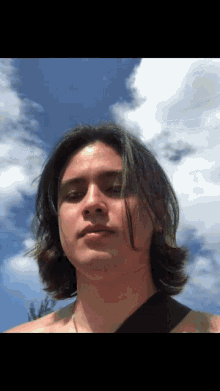 a man with long hair stands in front of a blue sky with clouds