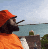 a man in an orange shirt is smoking a cigar in front of a body of water