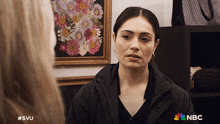 a woman in a black jacket is talking to another woman in front of a framed picture of flowers and a nbc logo
