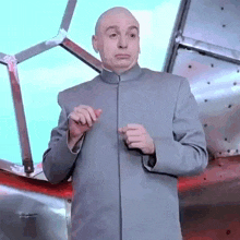 a bald man in a gray suit is making a funny face while standing in front of a window .