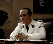 a man in a white suit is sitting at a desk and says shuffle mr. berner
