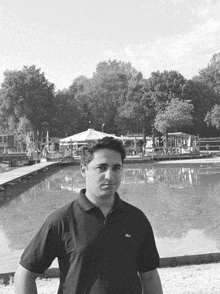 a man wearing a black lacoste shirt stands in front of a pond