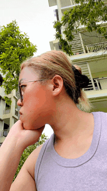 a woman wearing glasses and a purple tank top holds her hand to her chin in front of a building