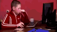 a young boy in a red jacket is sitting at a desk in front of a computer monitor .