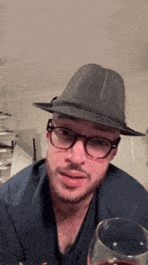 a man wearing glasses and a fedora holds a glass of wine