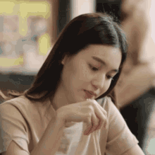a woman in a tan shirt is sitting at a table with her hand on her chin