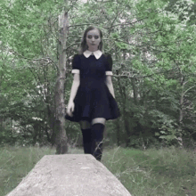 a woman in a black dress and knee high socks is standing on a stone in the woods .