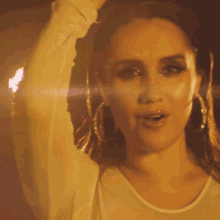 a woman wearing hoop earrings and a white shirt holds her hand to her forehead