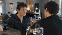 a man in a plaid shirt talks to another man in a black shirt