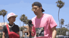 a man in a pink shirt holds a microphone in front of a boy wearing a white hat with the word supreme on it