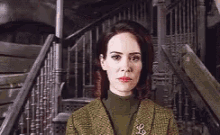 a woman in a green jacket and turtleneck is standing in front of stairs .