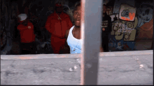 a man in a tank top stands in front of a wall with graffiti on it that says dg