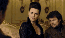 a woman in a black dress is sitting on a couch next to a man in a leather jacket .