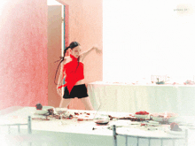 a woman in a red top and black shorts is standing in front of a messy table