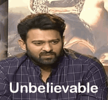 a man with a beard and a plaid shirt is sitting in front of a poster that says unbelievable .