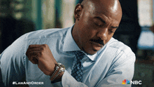 a man in a blue shirt and tie is looking at his watch with a nbc logo behind him
