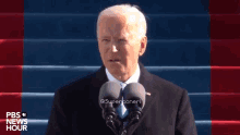 a man is giving a speech in front of two microphones and the words pbs news hour are on the bottom