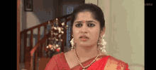 a woman in a red saree is sitting in front of a staircase and looking at the camera .