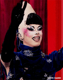 a drag queen wearing a blue top and black gloves holds her head