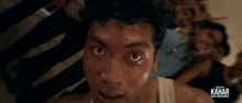 a man in a white tank top looks at the camera with a kahar logo behind him