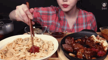 a woman in a plaid shirt is eating noodles and steak with chopsticks