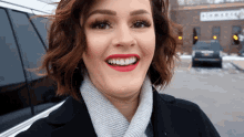 a woman wearing red lipstick and a scarf smiles in front of a car