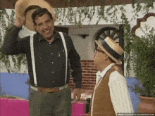 two men are standing next to each other and one is wearing a hat and suspenders