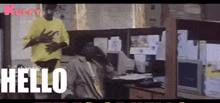 a man in a yellow shirt is standing next to a man sitting at a desk and says hello .
