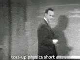a man in a suit and tie is standing in front of a blackboard and says toss-up physics