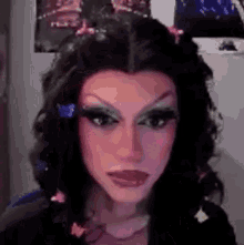 a drag queen with a wig and flowers in her hair is looking at the camera .