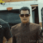 a man wearing sunglasses is standing in front of a sign that says ' ari watch '