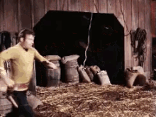 a man in a yellow shirt is standing in a barn with hay and barrels .