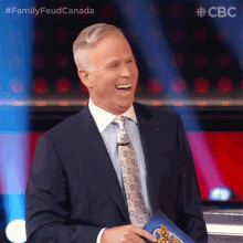 a man in a suit and tie is laughing while holding a clipboard with the words family feud canada written on it