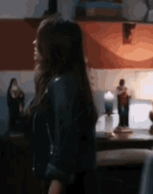 a woman in a black leather jacket is standing in front of a table with a lamp on it