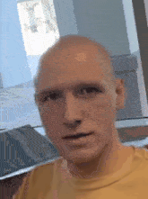 a bald man wearing a yellow shirt is standing in front of a window .