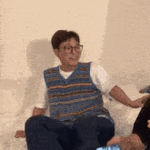 a man wearing glasses and a blue vest is sitting on a bed with his legs crossed .