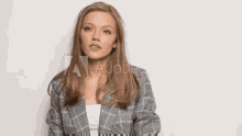 a woman in a plaid jacket is standing in front of a white wall with her hand on her chest .