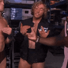 a shirtless man wearing a black jacket with the letter p on it talks to a group of people