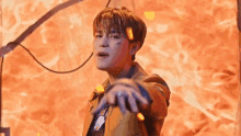 a young man in a brown jacket is standing in front of a fire and holding his hands out .