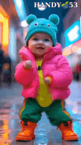 a baby wearing a pink jacket and a blue hat is standing in front of a sign that says " handy 53 "