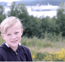 a young boy in a black jacket smiles in front of a large ship in the distance