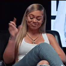 a woman in a white tank top and jeans is sitting in a chair with her legs crossed .