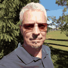 a man wearing sunglasses and a blue shirt stands in front of a pine tree