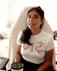 a woman in a white shirt with a dragon on it is sitting in a chair with a cup of coffee .